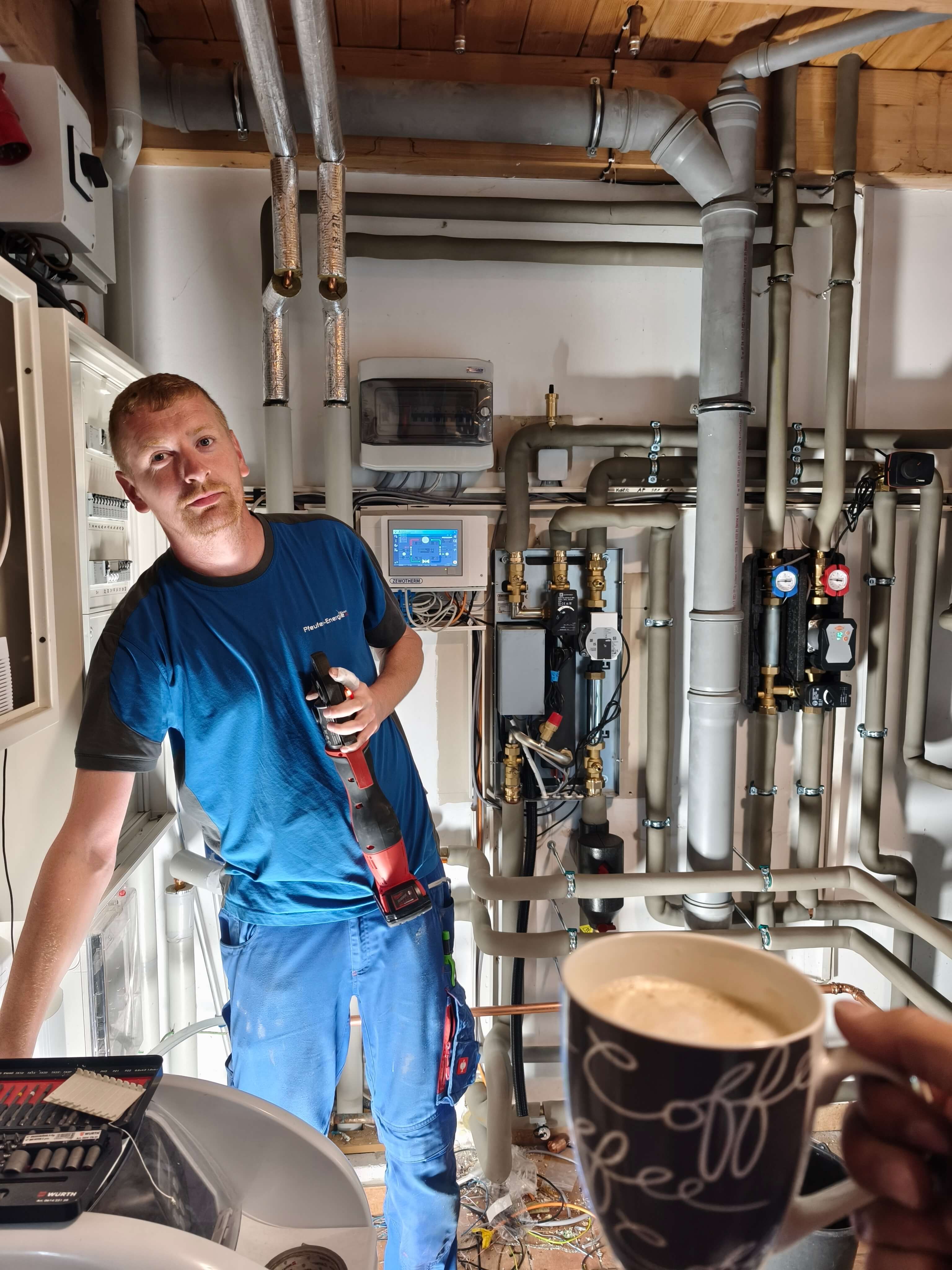 Ein Pfeufer-Energie Monteur in blauer Kleidung arbeitet an der Installation der Wärmepumpe in einem Technikraum, während er ein Werkzeug in der Hand hält.